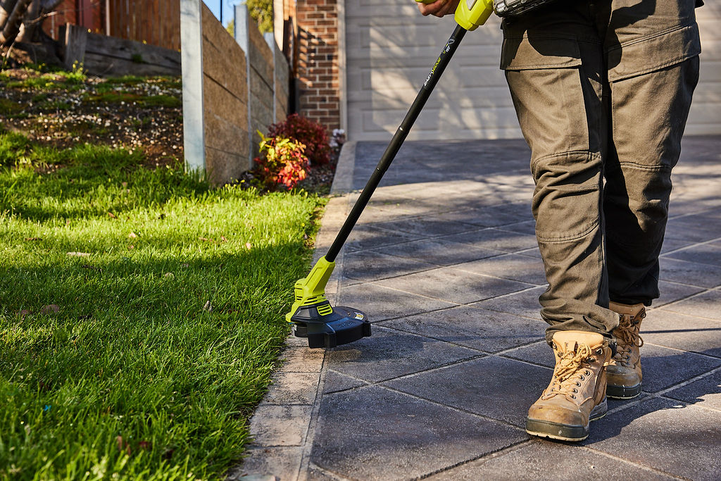 A man uses a RYOBI Line Trimmer on the grass growing at the edge of the driveway