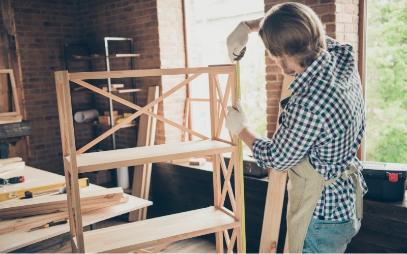 A man wearing a checked shirt and apron measures a panel on his DIY bookcase.