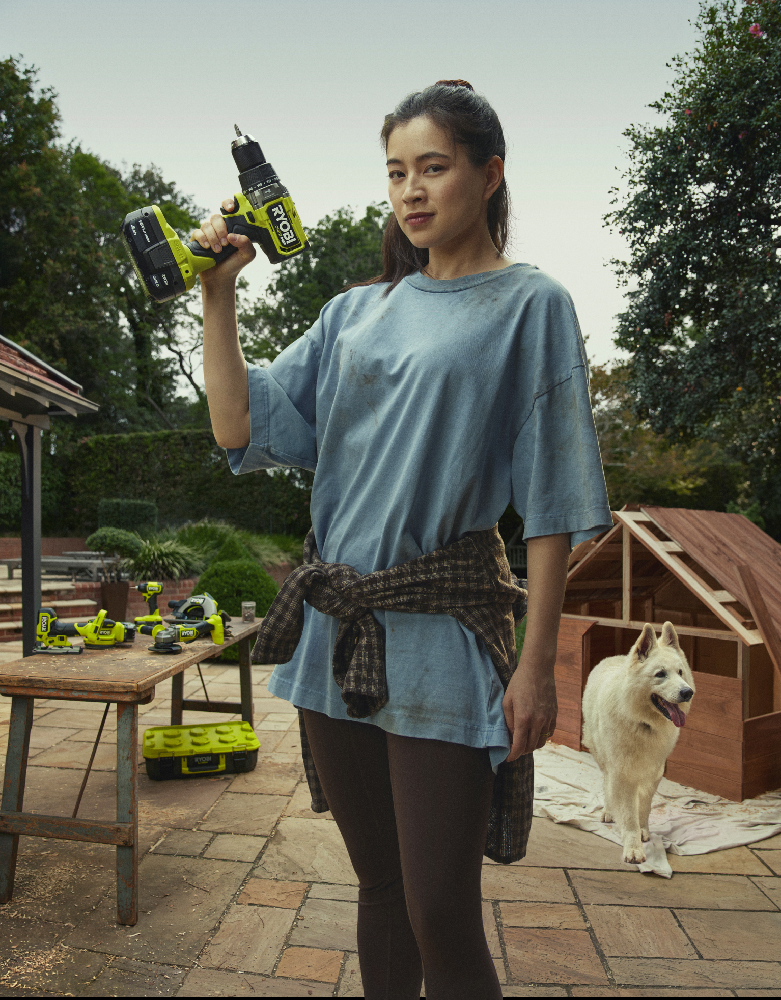 A woman stands holding a RYOBI Drill. Behind her is a DIY kennel, a fluffy white dog and a table full of RYOBI power tools. 